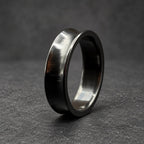 A black ring with a concave design and a polished finish. The ring is made of a dark material, possibly metal, and has a smooth, reflective surface. The ring is set against a wooden background.