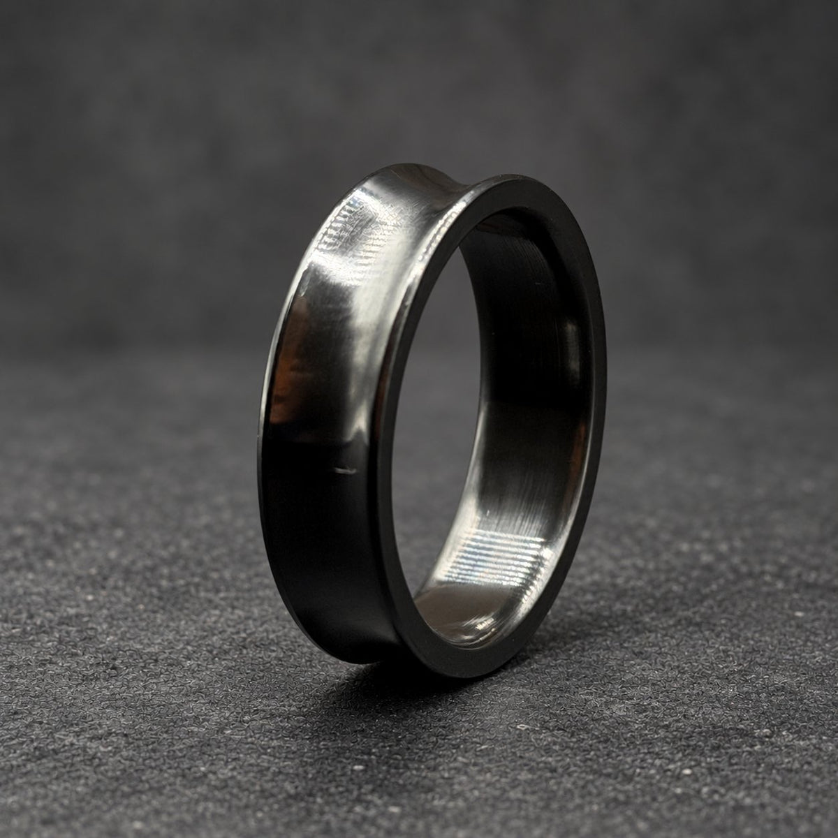 A black ring with a concave design and a polished finish. The ring is made of a dark material, possibly metal, and has a smooth, reflective surface. The ring is set against a wooden background.