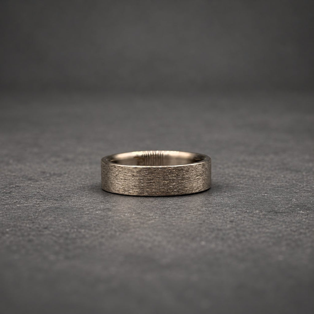 A flat band ring featuring a dark gray metallic finish with a distinctively textured, brushed surface. The ring has a uniform width and a smooth, polished interior edge, showcasing a simple, circular design with a matte appearance.