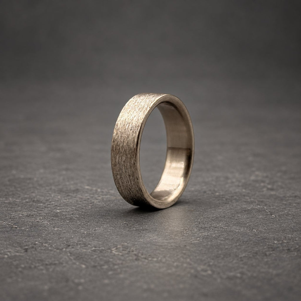 A silver-toned metal ring featuring a textured, brushed finish on the outer surface. The band has a consistent width and a smooth, polished interior, showcasing a metallic sheen that reflects light across its circular form.