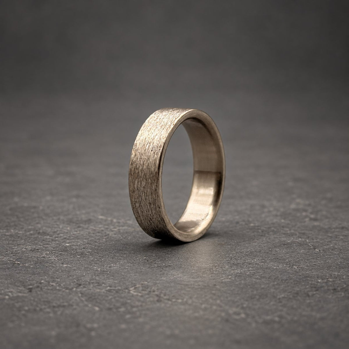 A silver-toned metal ring featuring a textured, brushed finish on the outer surface. The band has a consistent width and a smooth, polished interior, showcasing a metallic sheen that reflects light across its circular form.
