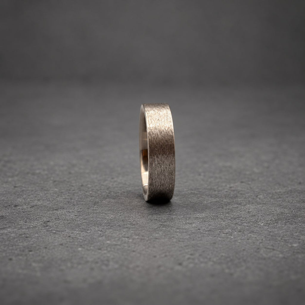 A dark gray metal ring with a brushed, textured finish. The band has a flat profile and a consistent width throughout, showcasing a matte appearance that reflects light subtly across its surface while standing upright on its edge.