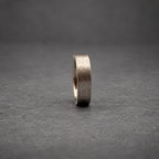 A dark gray metal ring with a brushed, textured finish. The band has a flat profile and a consistent width throughout, showcasing a matte appearance that reflects light subtly across its surface while standing upright on its edge.