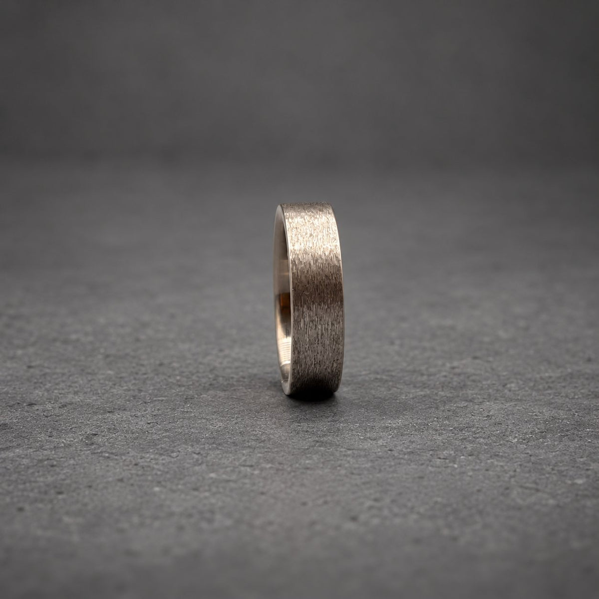 A dark gray metal ring with a brushed, textured finish. The band has a flat profile and a consistent width throughout, showcasing a matte appearance that reflects light subtly across its surface while standing upright on its edge.