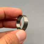 A circular ring with a dark gray, brushed metal exterior finish and a smooth, polished metallic interior surface. The band features a flat profile and a textured, matte appearance on the outer circumference of the metal ring.