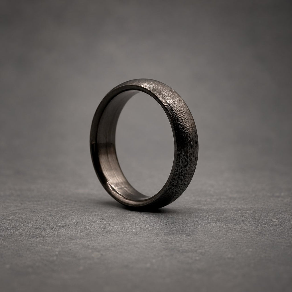 A solid black ring with a textured, matte exterior finish and a smooth, polished interior band. The circular band features a consistent width and a rounded profile, showcasing a dark, monochromatic aesthetic throughout the entire piece.