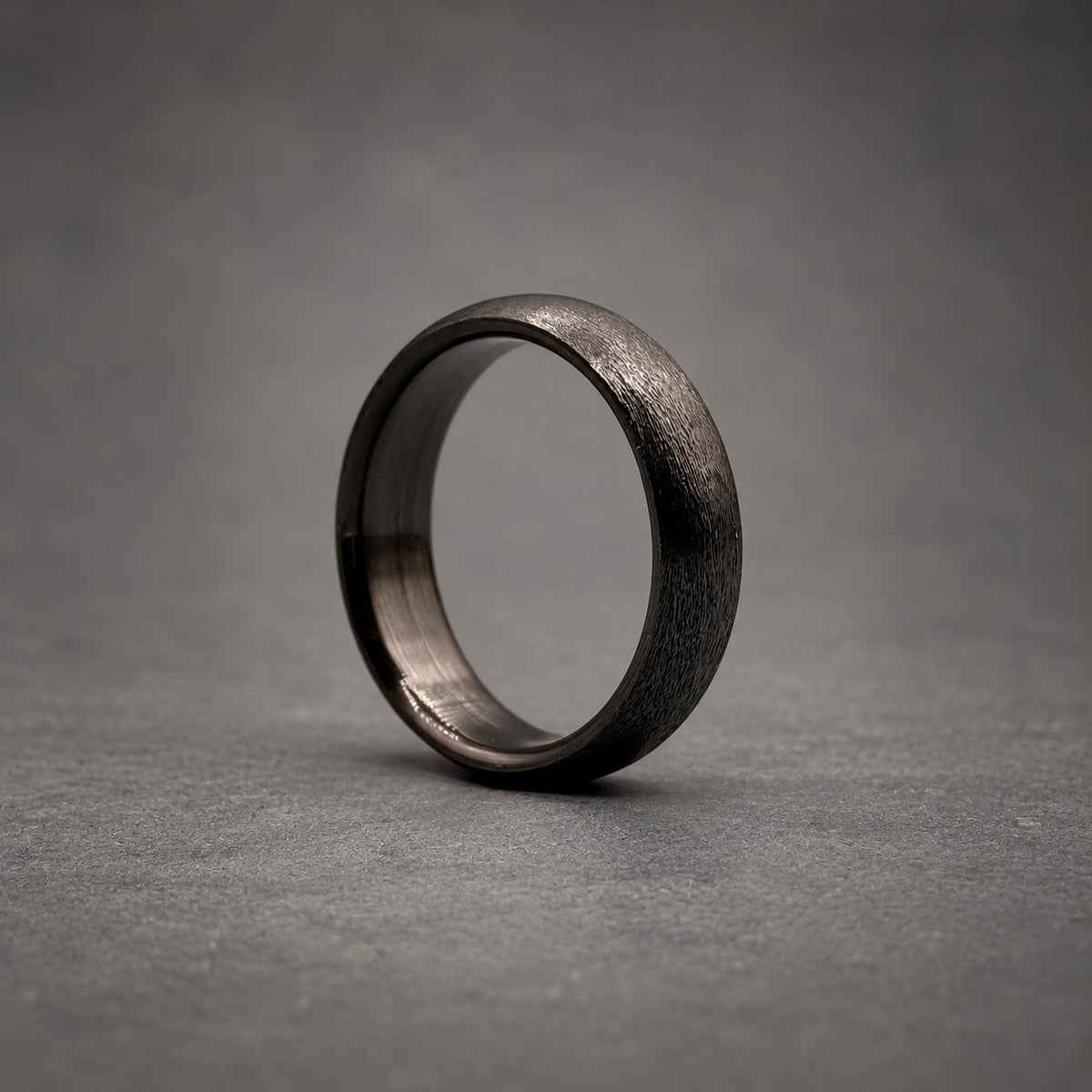 A solid black ring with a textured, matte exterior finish and a smooth, polished interior band. The circular band features a consistent width and a rounded profile, showcasing a dark, monochromatic aesthetic throughout the entire piece.