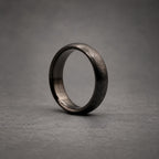 A solid black ring with a textured, matte exterior finish and a smooth, polished interior band. The circular band features a consistent width and a rounded profile, showcasing a dark, monochromatic aesthetic throughout the entire piece.