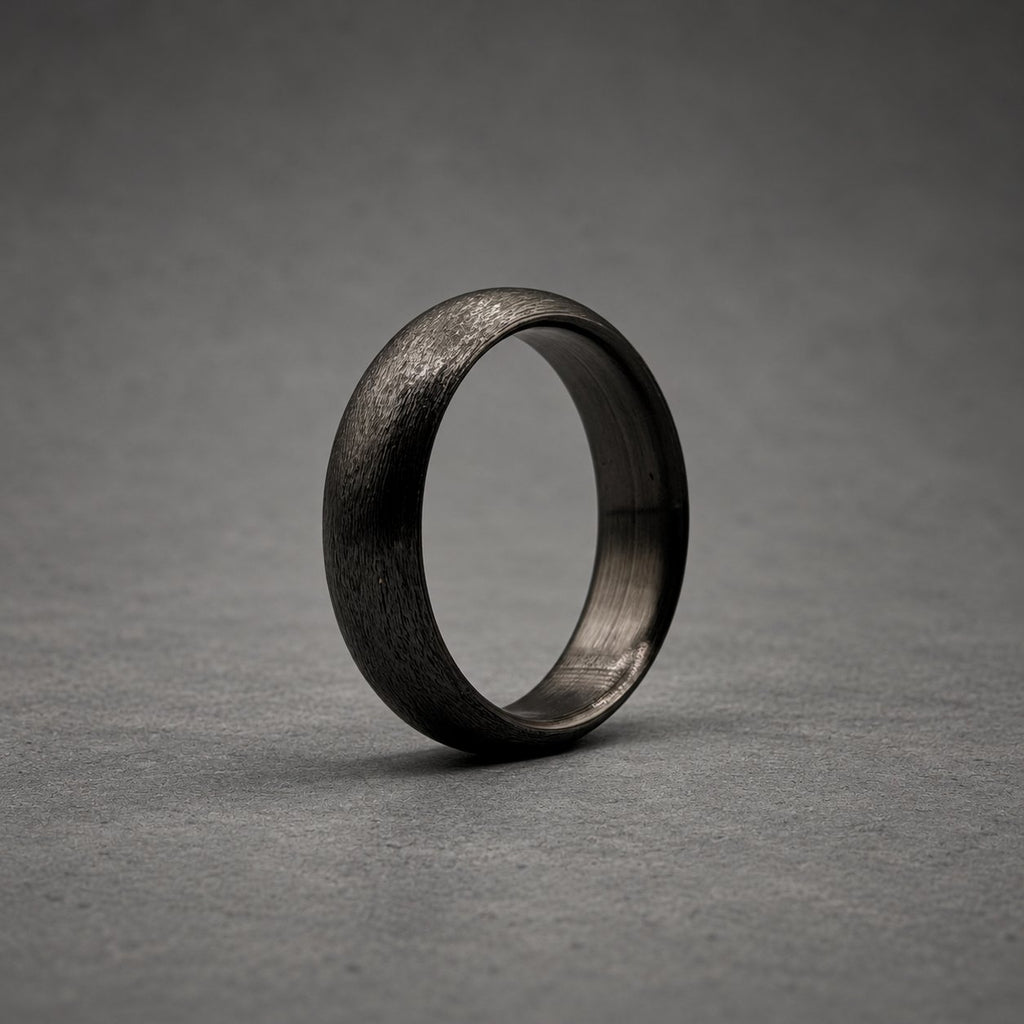 A solid black ring with a textured, matte exterior surface and a smooth, dark interior band. The ring features a rounded profile and a consistent dark charcoal color throughout, showcasing a simple, circular design without any additional embellishments or stones.