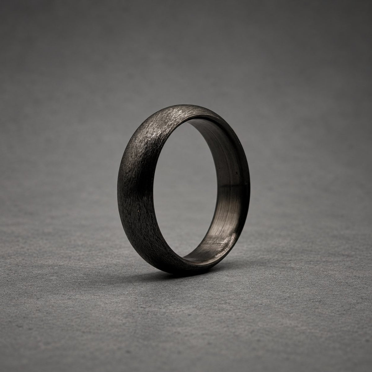 A solid black ring with a textured, matte exterior surface and a smooth, dark interior band. The ring features a rounded profile and a consistent dark charcoal color throughout, showcasing a simple, circular design without any additional embellishments or stones.