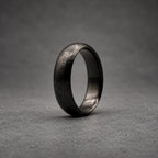 A dark gray ring with a textured, matte finish on the outer surface and a smooth, polished interior. The band has a rounded profile and a consistent width throughout, showcasing a deep charcoal color with subtle variations in the surface texture.