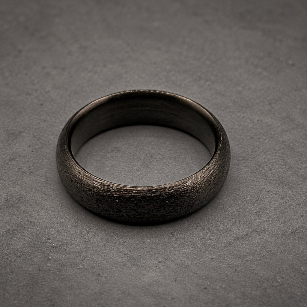 A circular ring with a dark charcoal gray color and a textured, matte finish. The band has a consistent width and a smooth, polished interior surface that contrasts with the rough, granular appearance of the outer circumference.