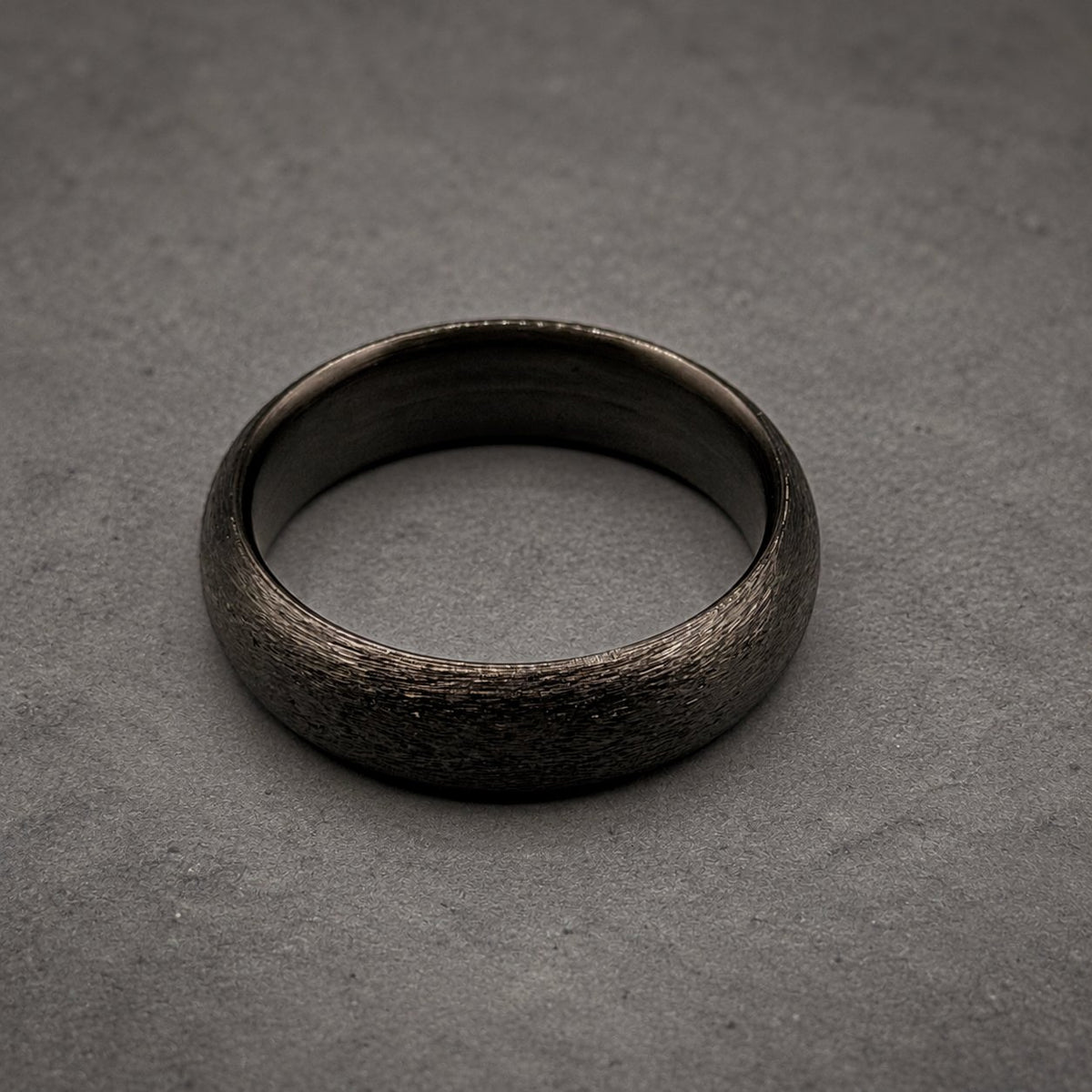 A circular ring with a dark charcoal gray color and a textured, matte finish. The band has a consistent width and a smooth, polished interior surface that contrasts with the rough, granular appearance of the outer circumference.