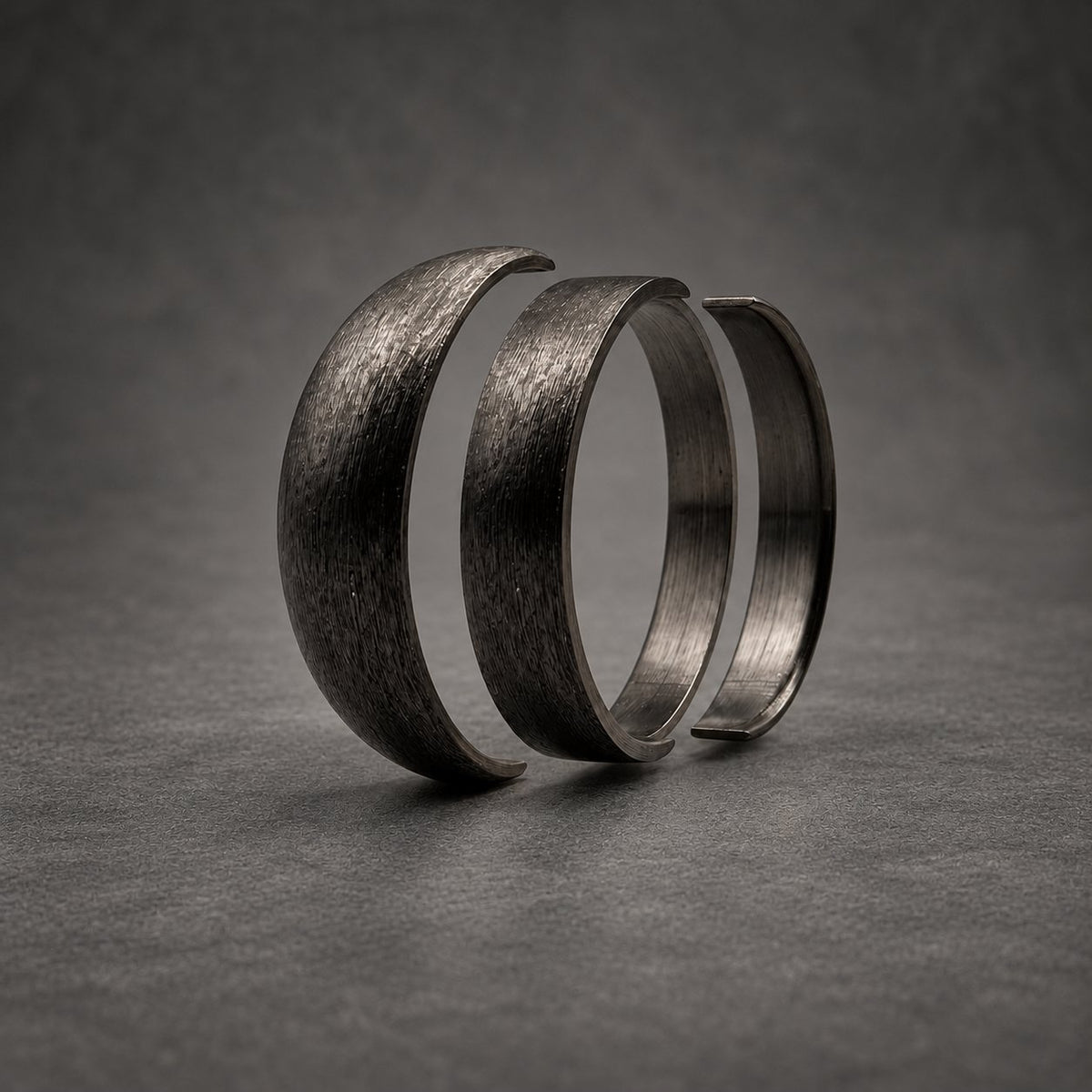 Three dark gray rings arranged in a row, each featuring a textured, brushed metal finish. The rings are positioned vertically, showing a consistent matte appearance across their curved surfaces, creating a layered visual effect.
